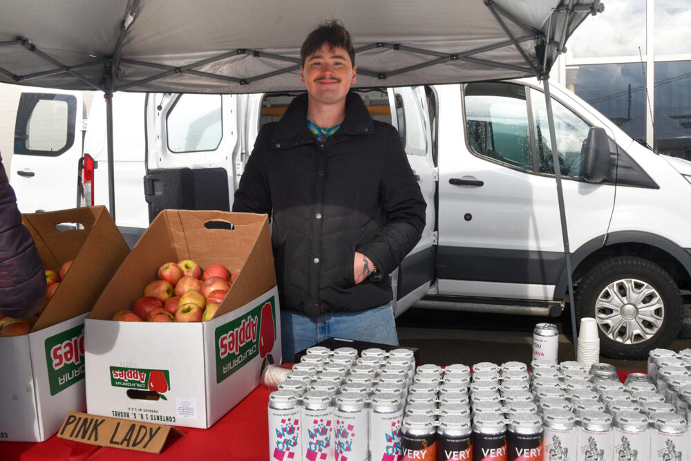 Joey stands at Far West Cider & Chinchiolo Family Farms' stand at the Ferry Plaza Farmers Market. Their table holds boxes of apples and rows of hard cider cans.