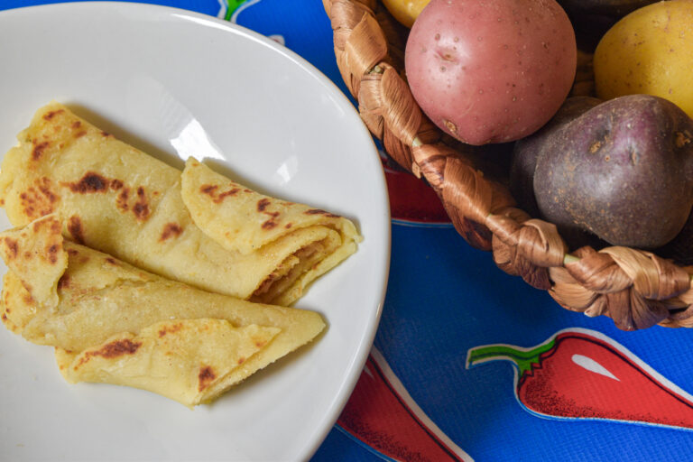 Two rolled potato pancakes on a white dish, next to a basked of multicolor potatoes