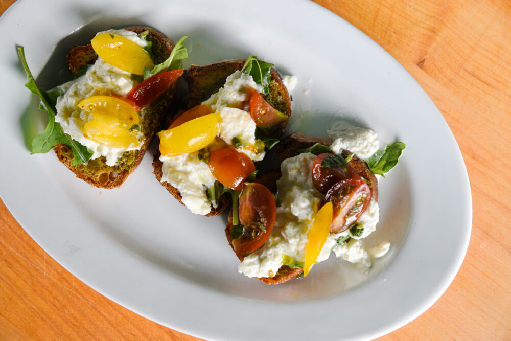 Bruschetta with stracciatella, pesto, and tomatoes, served on a white dish on a wooden board