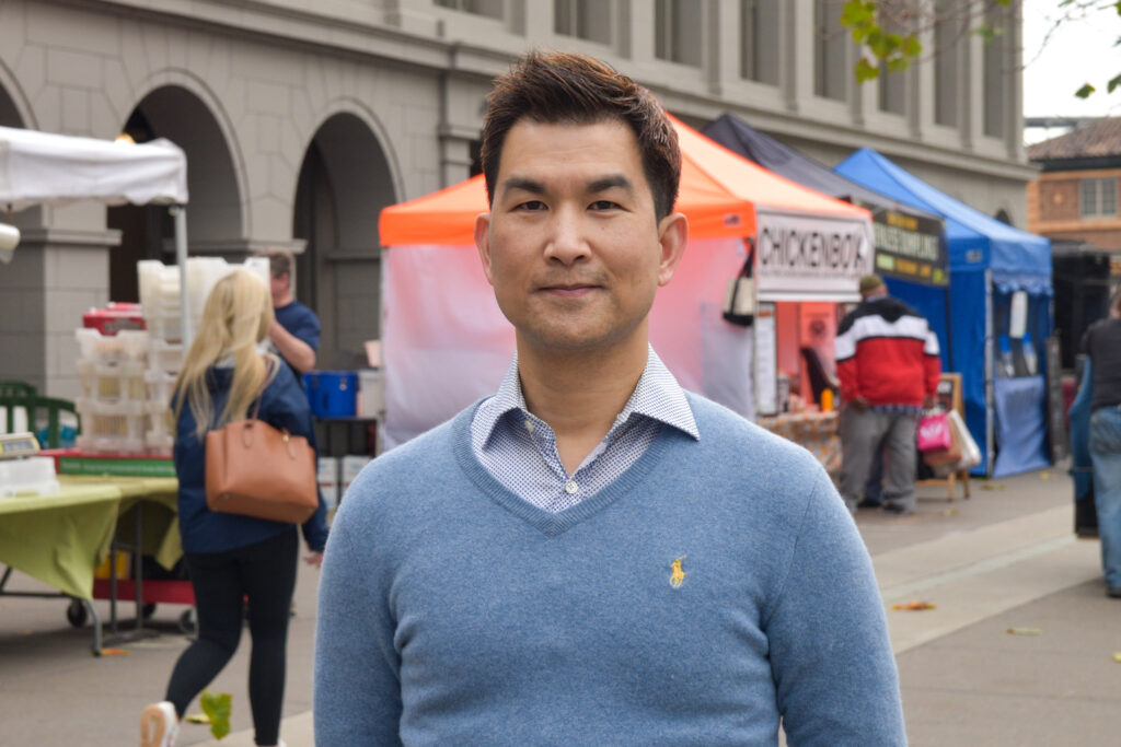 Foodwise volunteer, Wayne. poses for a photo in front of stands at the Ferry Plaza Farmers Market.